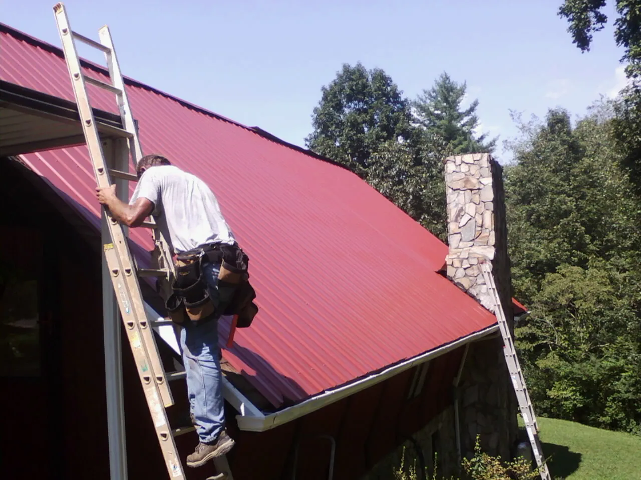 Roofer on a ladder beside a red metal roof and stone chimney — Soffit & Fascia Repair by Roofing Brothers in East Alton